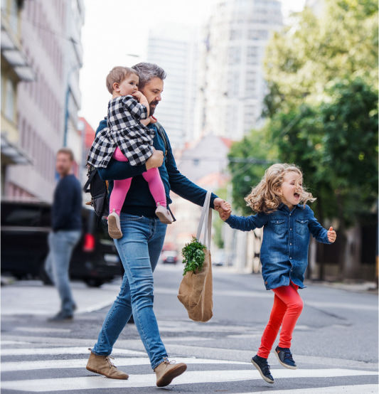 Father crossing the street carrying a baby while holding groceries and his young daughter's hand as she skips