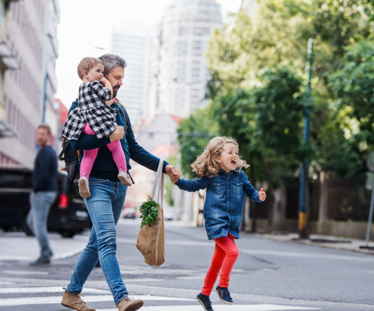 Father crossing the street carrying a baby while holding groceries and his young daughter's hand as she skips