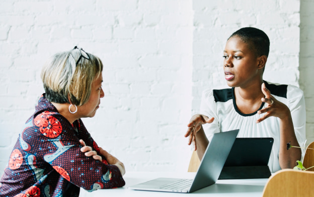 Two women sitting at a table and conversing. One woman has a laptop in front of her as they talk.