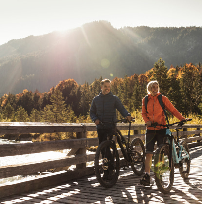 A man and a woman standing next to their bikes, holding the handlebars, smiling and looking into the distance.