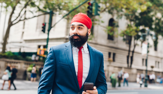 A man standing on a city street, holding a phone while looking in the distance.