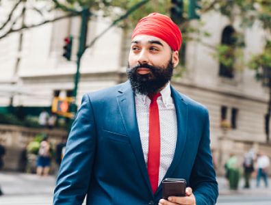 A man standing on a city street, holding a phone while looking in the distance.