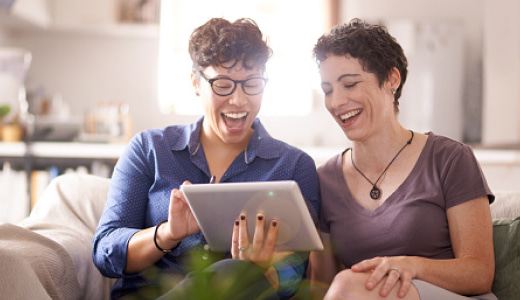 Two women sitting on a couch looking at a tablet, and laughing.