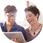 Two women sitting on a couch looking at a tablet, and laughing.