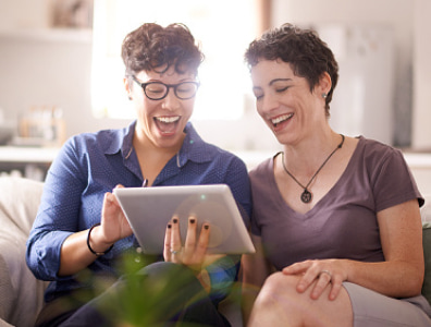 Two women sitting on a couch looking at a tablet, and laughing.