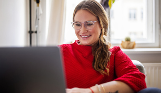 A woman sitting in an apartment, smiling while using a laptop.