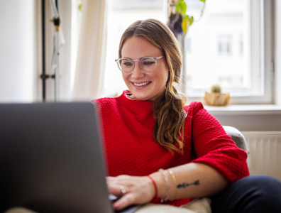 A woman sitting in an apartment, smiling while using a laptop.