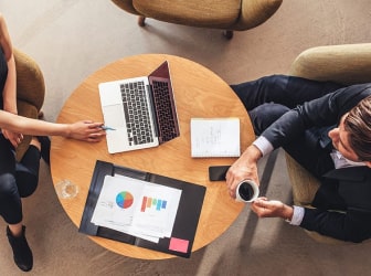 A bird's-eye view of two people sitting at a small table with a notebook and report. One person is holding a cup of coffee while the other points to a laptop screen.
