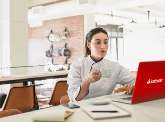 A woman sitting at a table in an office looking at a Santander-branded laptop. She is holding a mug in her right hand. On the table in front and to the left of her are a saucer, two notebooks, a pen, and a smartphone.