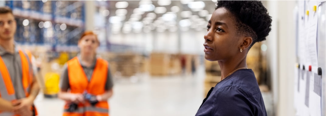 A woman standing near a wall in a warehouse, looking into the distance. There is a clipboard behind her and two workers in orange vests to her left.