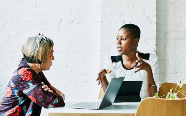 Two women sitting at a table having a conversation. Both have their laptops open in front of them.