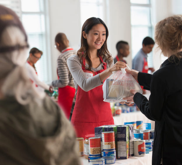 A woman in a red apron handing a plastic bag of canned food to another woman who is facing away from the camera. There are other people in red aprons in the background dealing with canned goods.