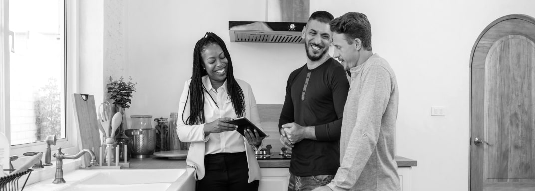 A woman and two men standing in the kitchen, smiling brightly. The woman is holding a tablet in both hands and tilting the screen toward the men.