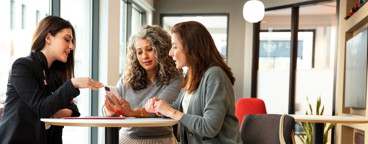 A female Santander employee sitting at a table with two women, looking at a smartphone screen.