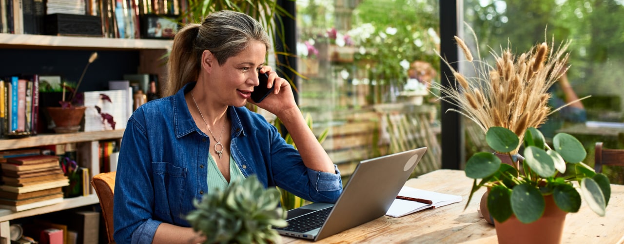 A woman sitting at a desk behind an open laptop and a notebook. The woman is smiling while talking on her smartphone.