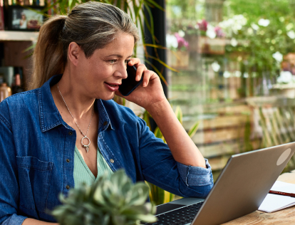 A woman sitting at a desk behind an open laptop and a notebook. The woman is smiling while talking on her smartphone.