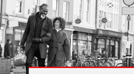 A man and woman, locking arms, walking through a city street while smiling at each other.