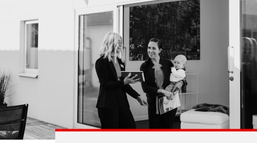 A female employee holding a tablet screen toward another woman holding a baby in her arms. Both are smiling.