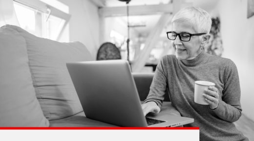 An older woman with a coffee mug in one hand, sitting down and looking at a laptop screen.