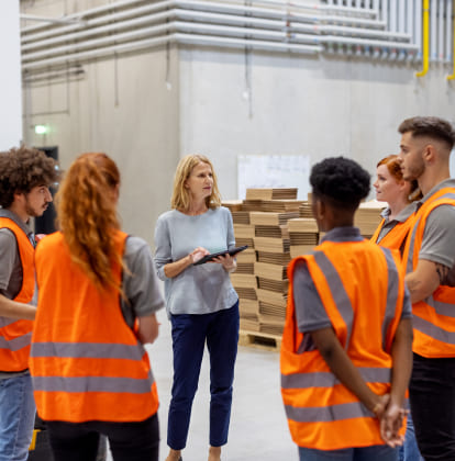 A woman with a tablet talking to a group of 5 workers in bright orange vests in a warehouse.