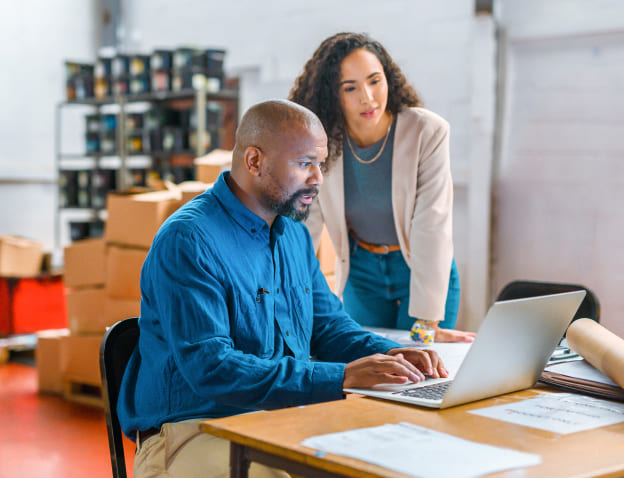 A man sitting at a table and a woman standing over his shoulder, both gazing at a laptop screen in a warehouse office setting.