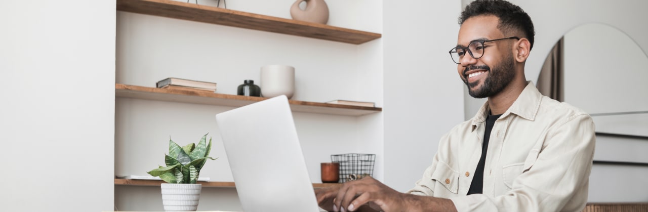 A man sitting at a table, looking at his laptop and smiling.