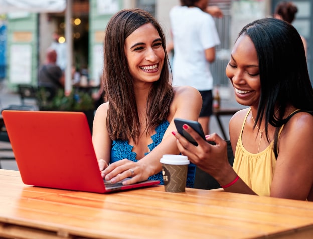 Two women sitting at a table, one with a laptop in front of her and the other with her phone in her hand. Both are smiling.