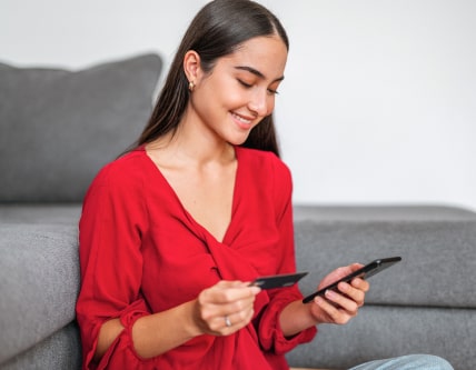 A woman sitting down holding a credit or debit card in one hand and her phone in the other, looking at the screen and smiling.