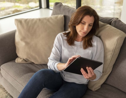 A woman sitting on a couch looking at the screen of a tablet.