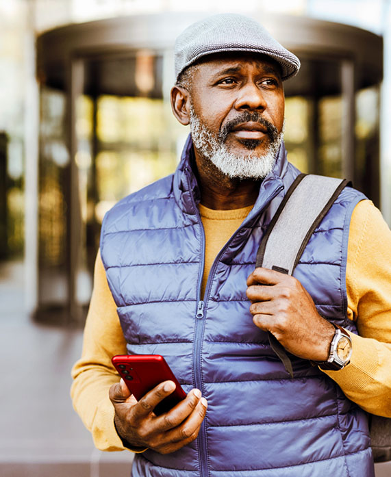 Man with hat and vest walking outside looking away phone