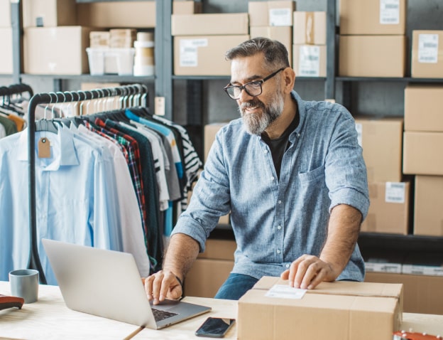 A man in a button-down shirt and jeans, sitting in a business office, typing on a laptop. There are two clothes racks of shirts and a wall of shelves full of boxes in the background.