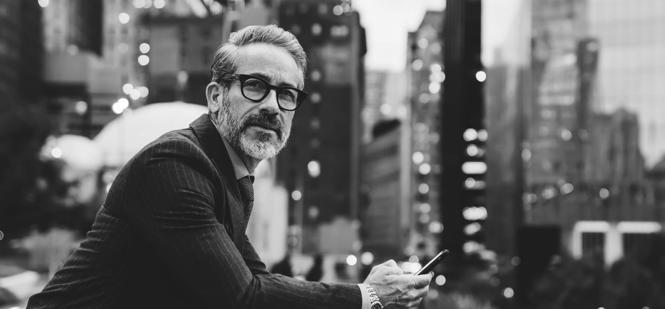 A man holding a smartphone in his hands and leaning against a railing outside, with skyscrapers in the background. He is staring toward the camera.