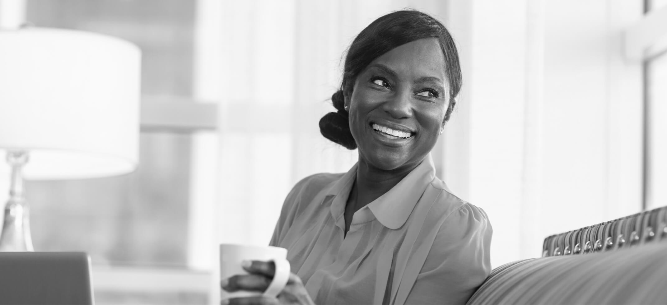 A woman with a coffee mug in hand, sitting in a room and smiling while looking off to the side.