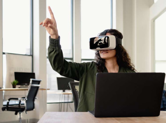 A woman wearing a VR headset sitting in front of a laptop and pointing.