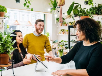 A man and woman in a plant shop standing at a counter speaking to the shop owner, who is pointing to a tablet.