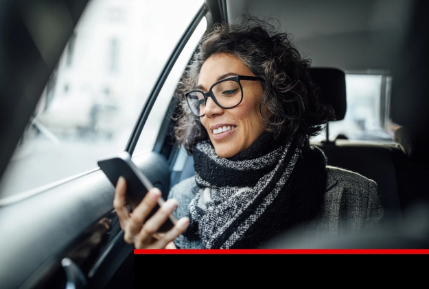 A woman in glasses and scarf checks her phone in the passenger seat of a car