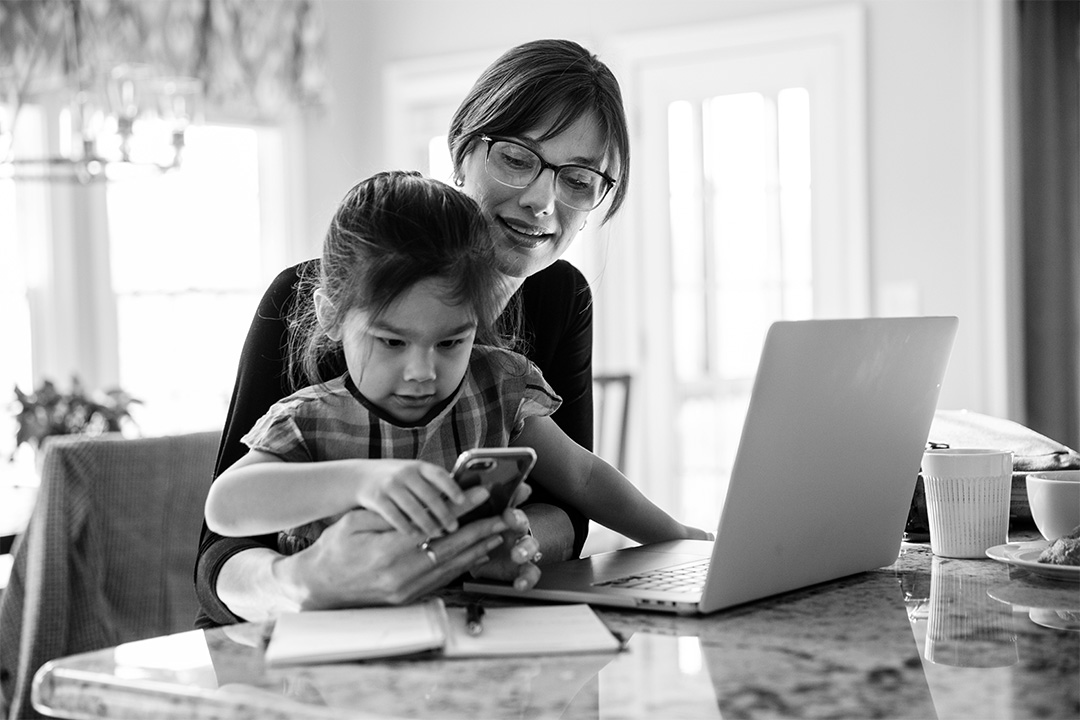 Daughter sitting on a mother's lap playing with a cell phone, while mom works on a laptop.